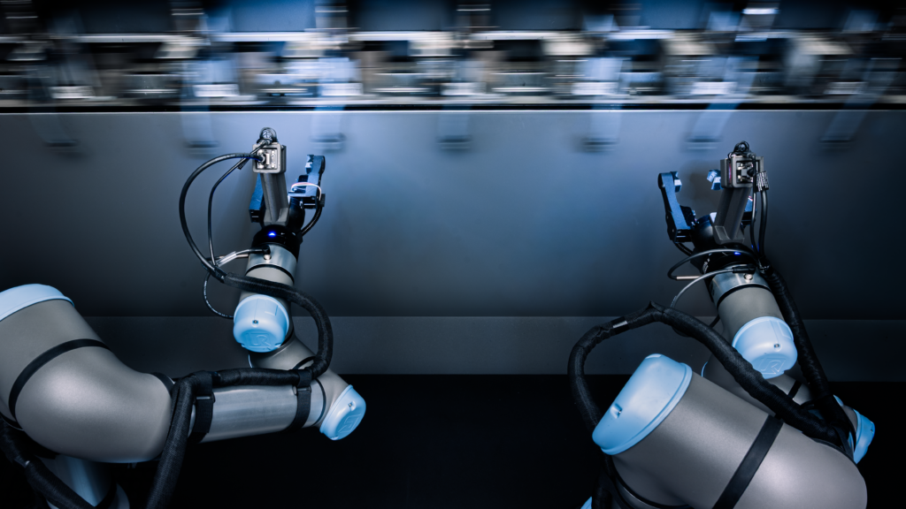Two robotic arms equipped with tools operate in sync on a manufacturing production line, with blurred machine parts moving above them in the background.