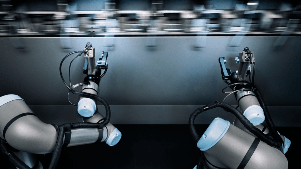 Two robotic arms with cables and sensors work together in a factory setting, positioned under a blurred, fast-moving conveyor belt or assembly line, suggesting automation and advanced manufacturing processes.