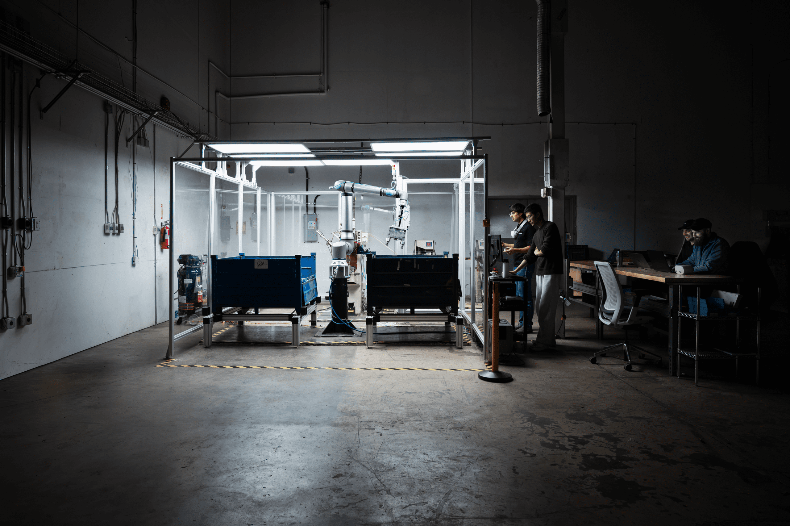 A dimly lit industrial warehouse shows two people monitoring a robotic arm working between large bins, with desks and computers to the right and utility pipes on the wall to the left.