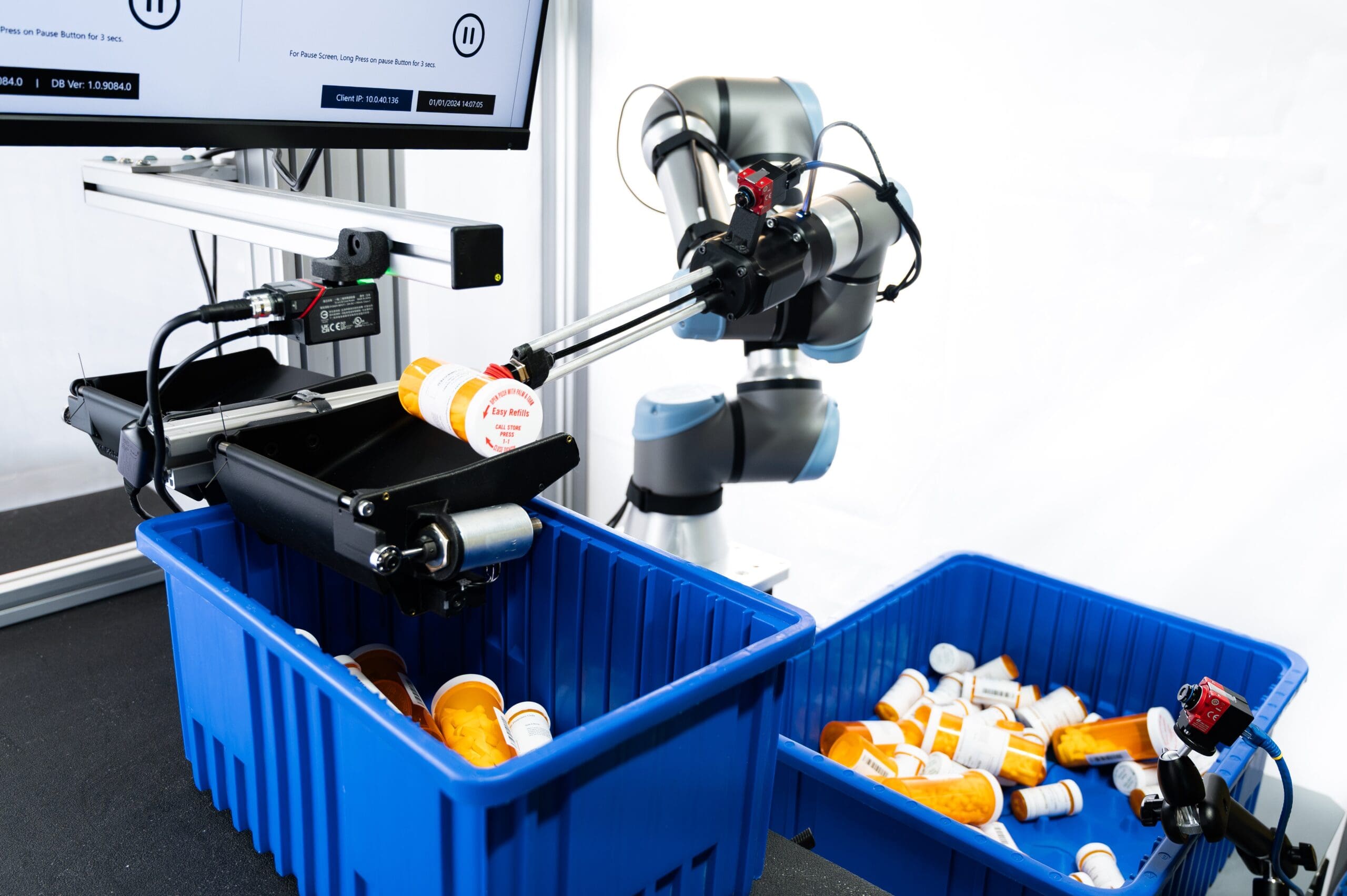 A robot arm sorts prescription pill bottles, moving them from a conveyor belt into blue bins. A computer monitor and sensors are visible in the background.