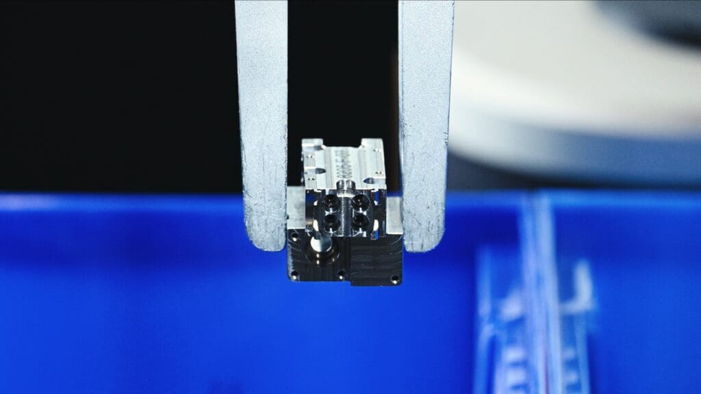 A robotic arm holds a small metallic component above a blue container, possibly in an industrial or manufacturing setting.