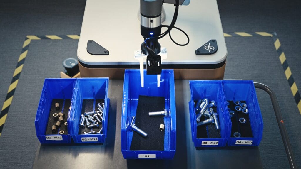 A robotic arm hovers over blue bins filled with metal bolts and nuts, labeled by size, on a workstation with a gray surface and safety tape marking the area.