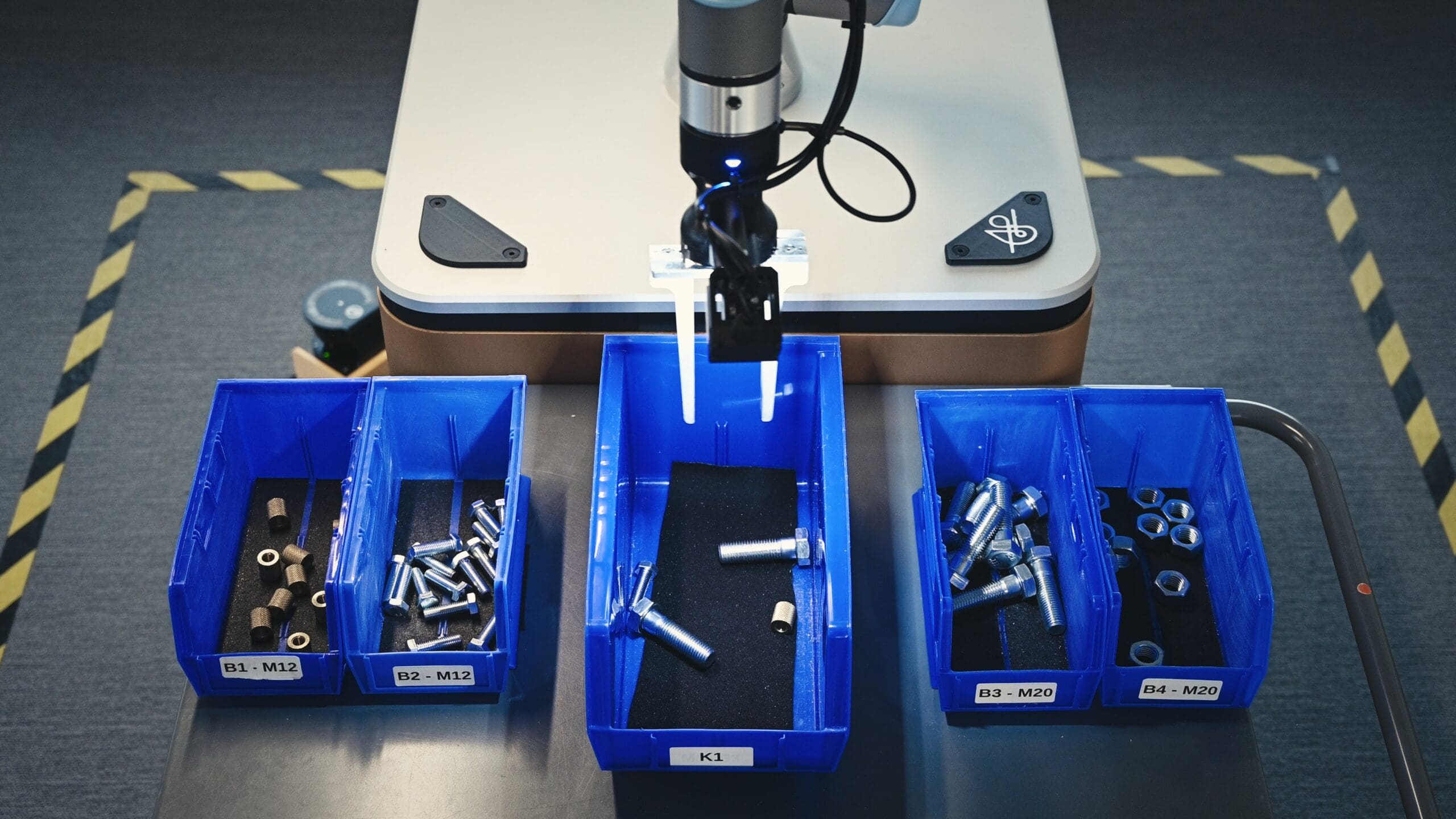 A robotic arm hovers over blue bins filled with metal bolts and nuts, labeled by size, on a workstation with a gray surface and safety tape marking the area.