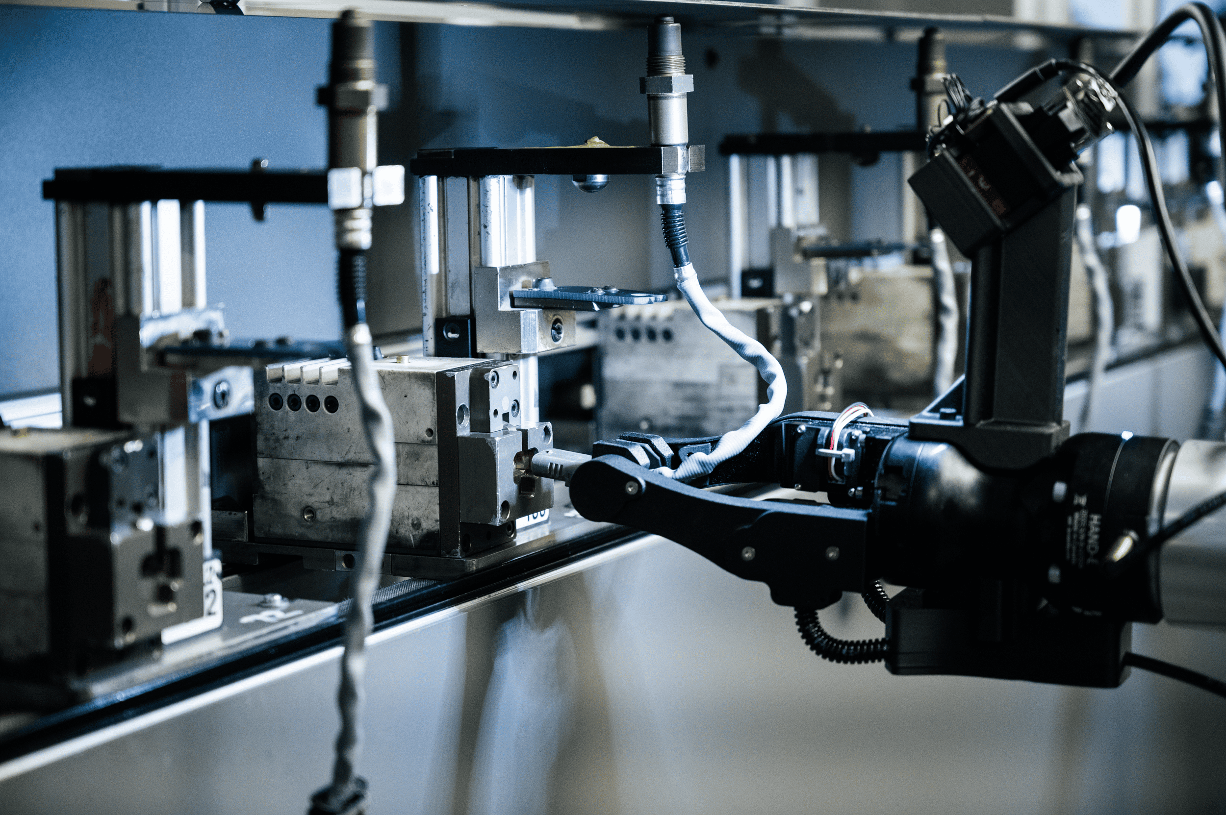 A robotic arm with a gripper is operating in an industrial setting, interacting with metal machinery components lined up on a workbench.