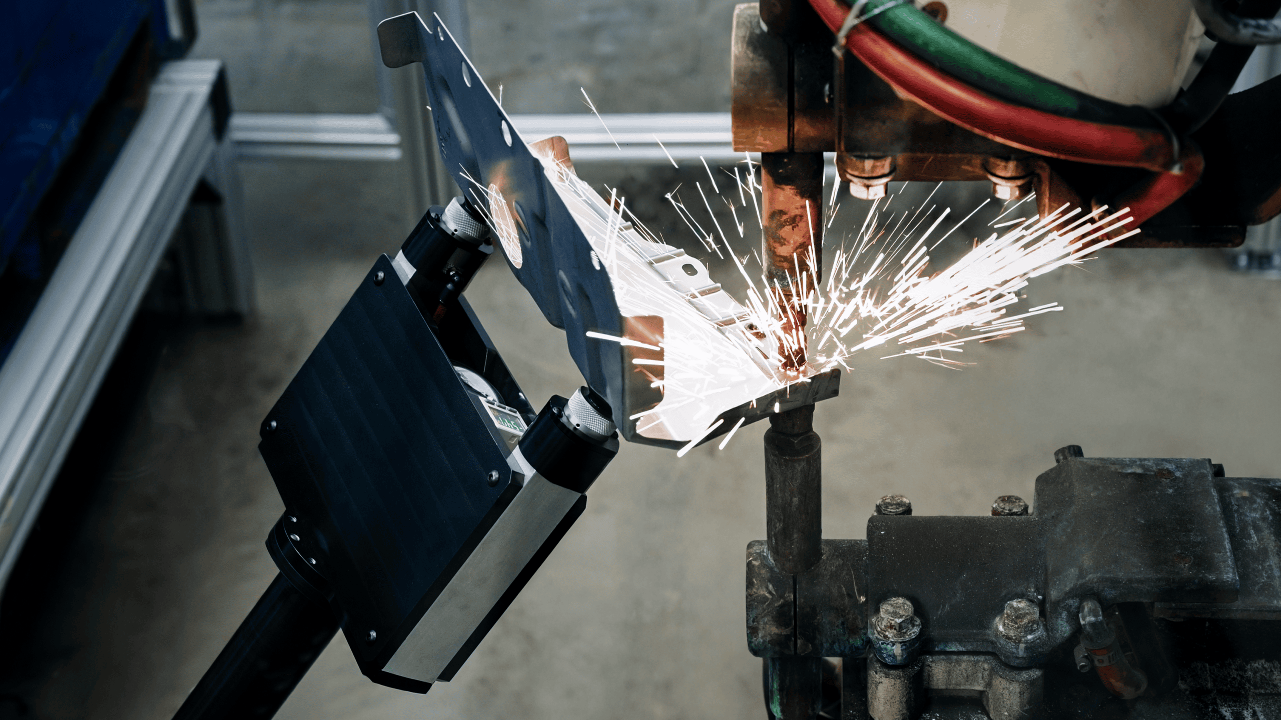 Close-up of a machine welding a metal piece, with bright sparks flying. The industrial setting shows robotic equipment and cables, highlighting automated manufacturing.